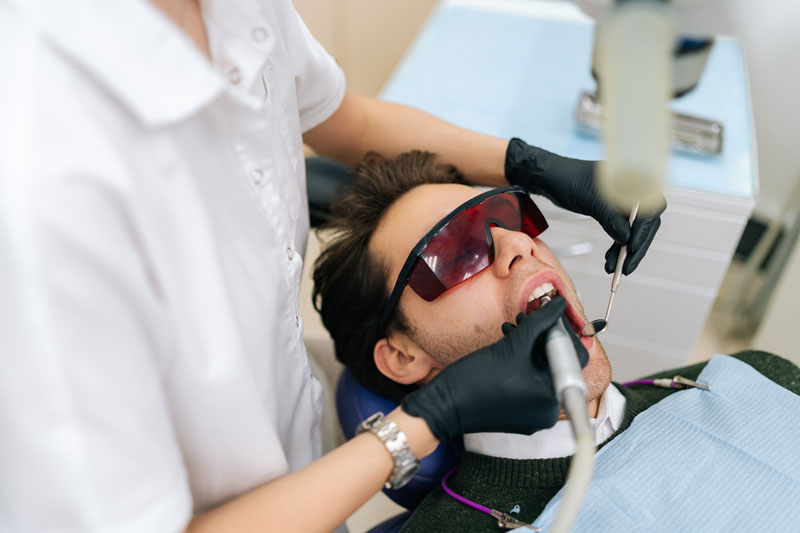 Female dentist using modern diode dental laser for periodontal care, top view. Male patient wearing protective glasses, preventing eyesight damage.