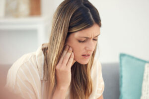Woman sitting on a couch holding her jaw in pain, possibly experiencing a toothache or dental discomfort.