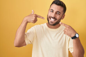 Smiling man pointing at his bright white teeth, wearing a casual t-shirt, standing against a vibrant yellow background.